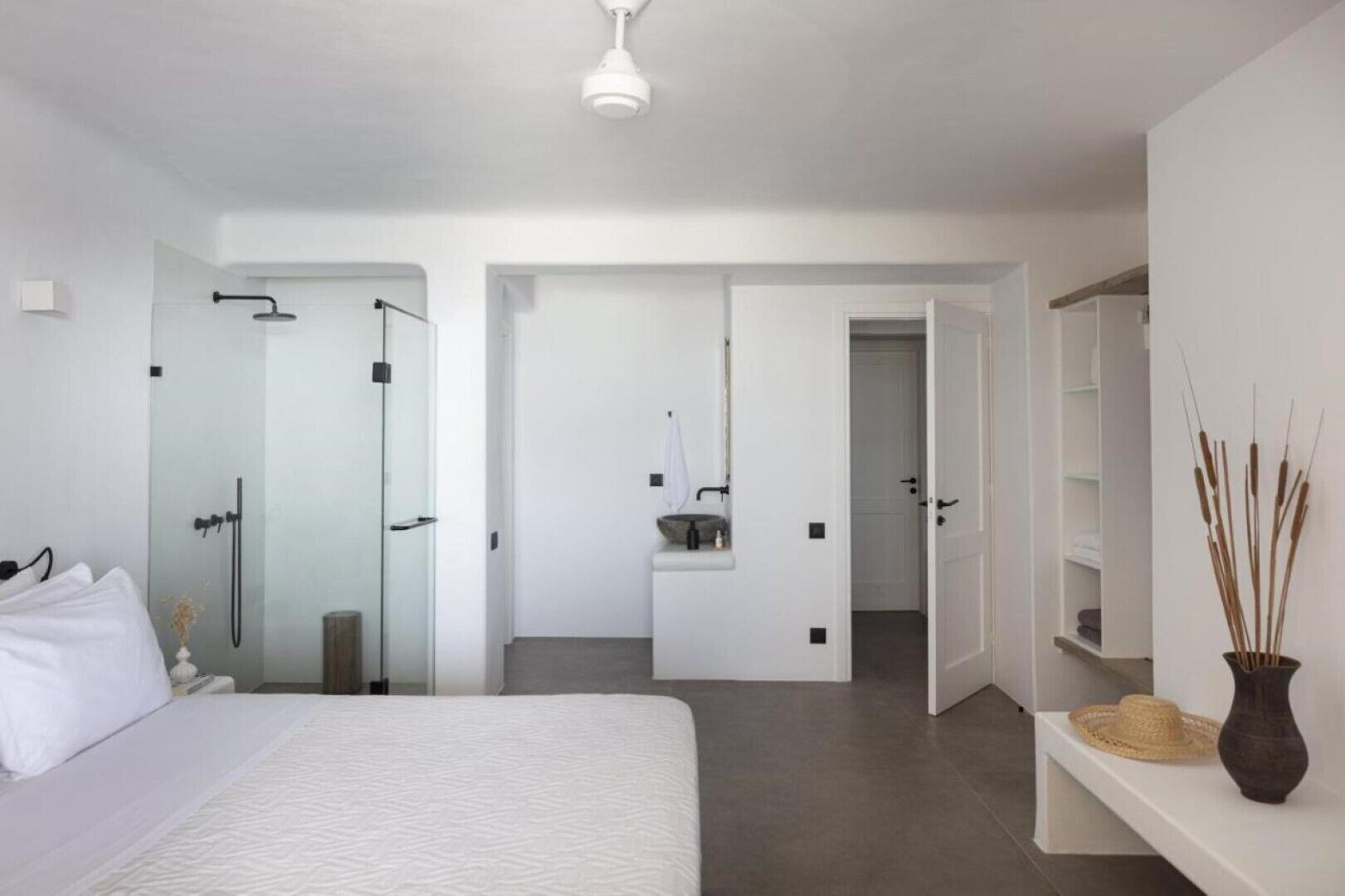 Ein modernes Schlafzimmer mit einem großen Bett, einer Glasdusche, einem Schminktisch mit Spiegel und einer Vase mit Schilf auf einem Regal.