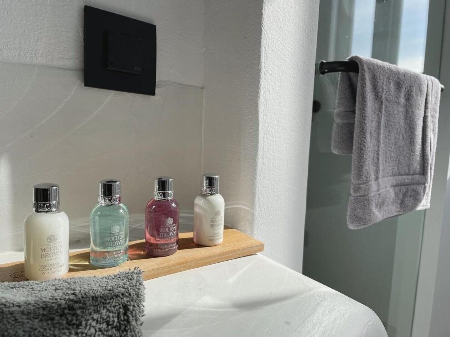 Un baño con un lavamanos blanco, un estante de madera con cuatro botellas de artículos de aseo, una toalla gris colgada en un perchero y un enchufe eléctrico negro en la pared.