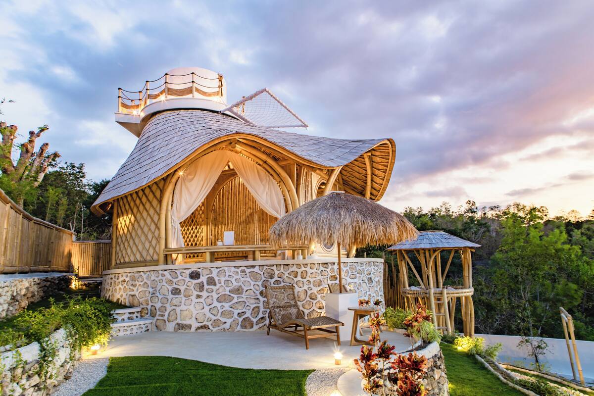 A unique, dome-shaped building with a stone exterior and a thatched roof, featuring a seating area with a table and chairs, surrounded by a well-maintained garden.