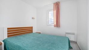 A bedroom with a bed, a nightstand, a lamp, a window with curtains, and a radiator. - Apartment with a balcony, 300 meters from the beach (Agde)