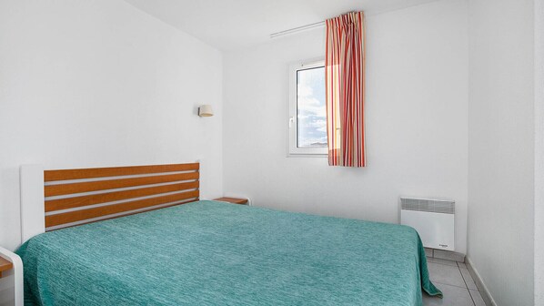A bedroom with a bed, a nightstand, a lamp, a window with curtains, and a radiator. - Apartment with a balcony, 300 meters from the beach (Agde)