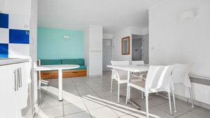 A modern, well-lit room with a white table and chairs, a teal-colored sofa, and a small kitchenette area. - Apartment with a balcony, 300 meters from the beach (Agde)