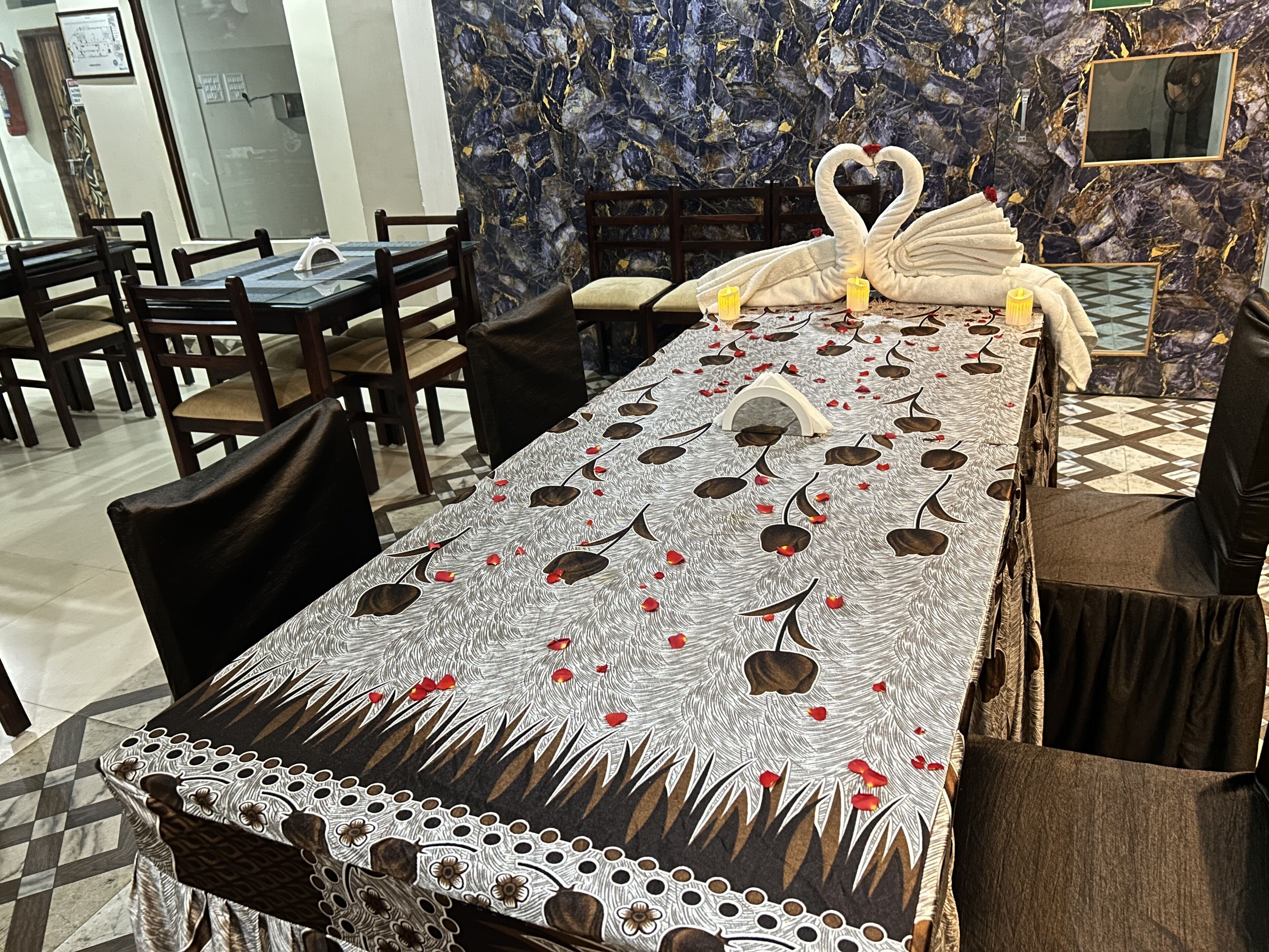A dining area with a long table set for two, featuring a decorative table runner with a leaf pattern and a swan centerpiece.
