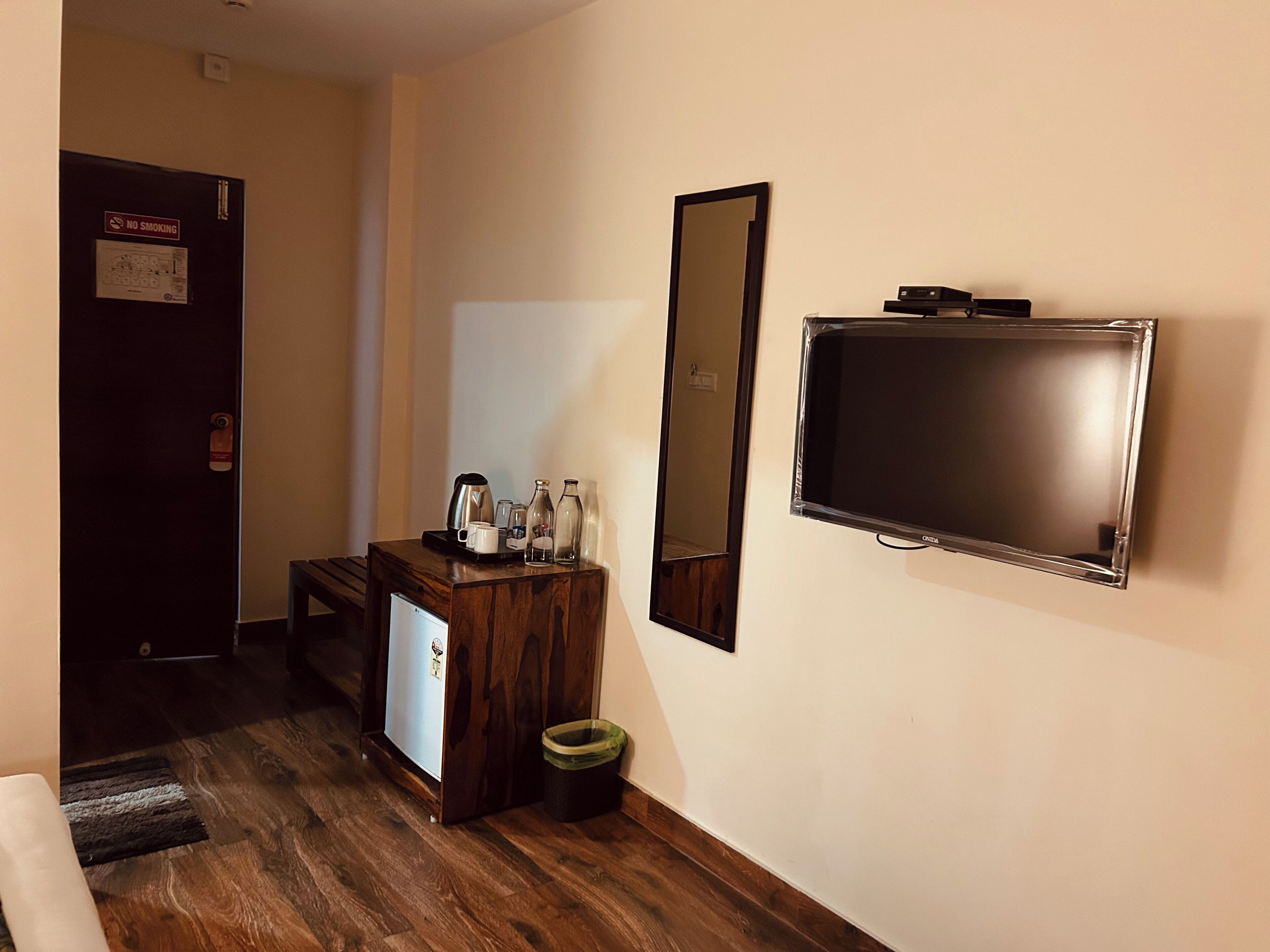 A hotel room with a wooden desk, a television, a mirror, a trash bin, and a coffee machine.
