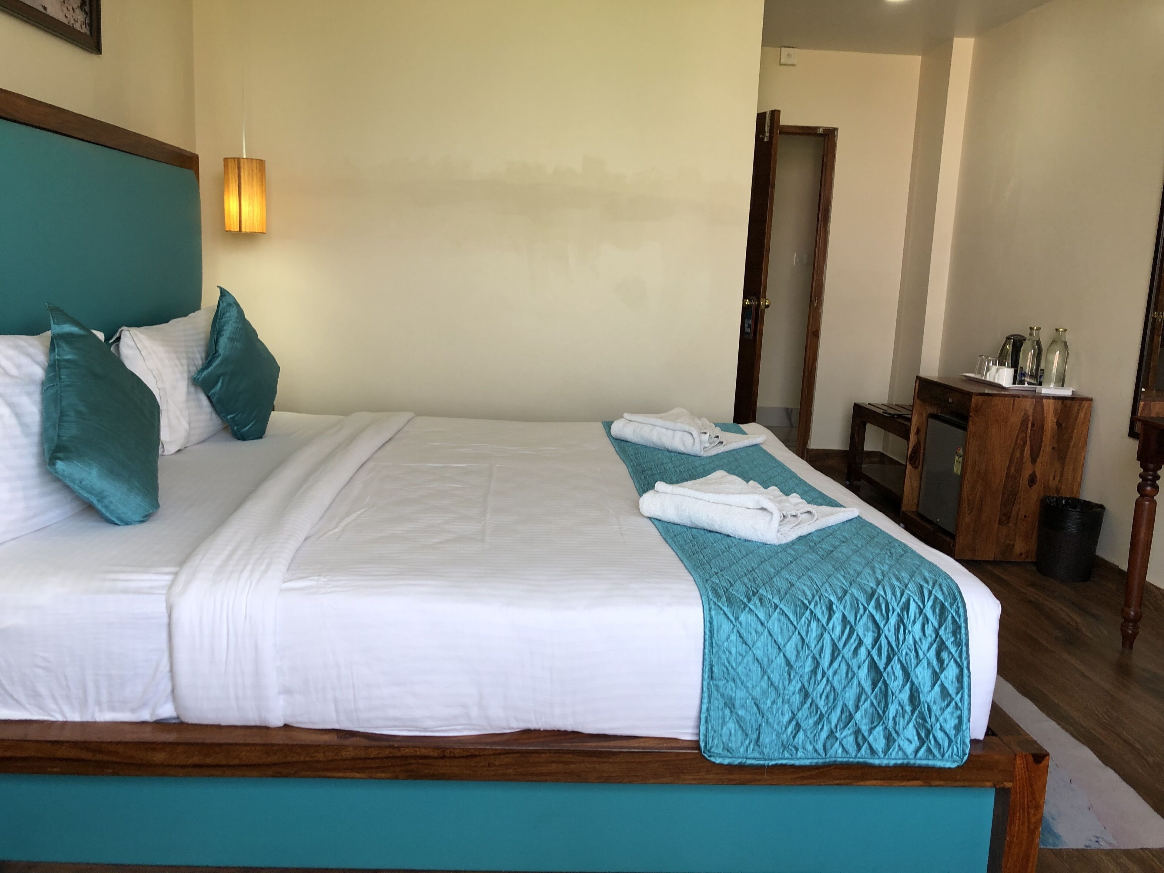 A neatly made bed with white linens and teal pillows, a teal quilted throw blanket, and two folded towels on the bed.