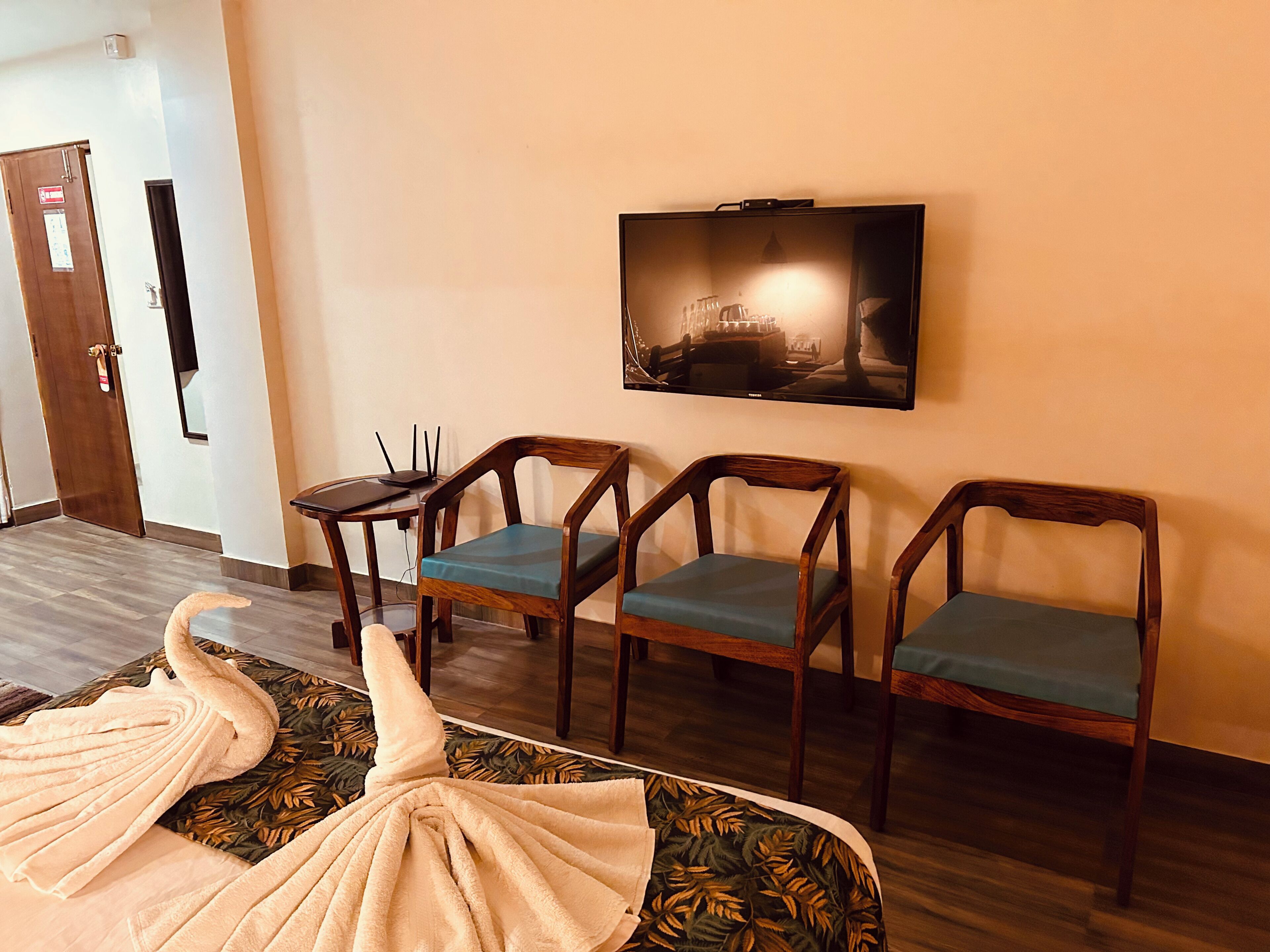 A hotel room with a flat-screen TV mounted on the wall, a sofa with a floral pattern, and three wooden chairs with green cushions.