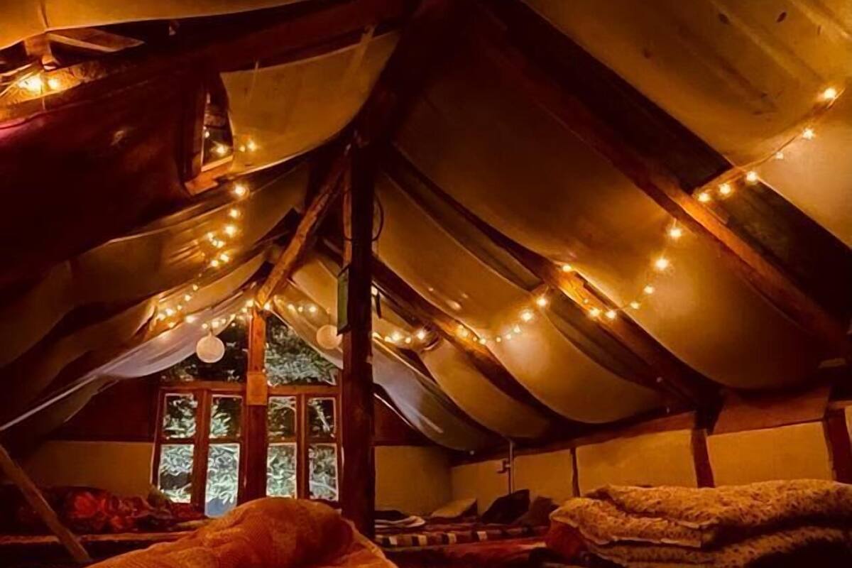A cozy attic bedroom with a sloped ceiling, string lights, and a window with a view of greenery.
