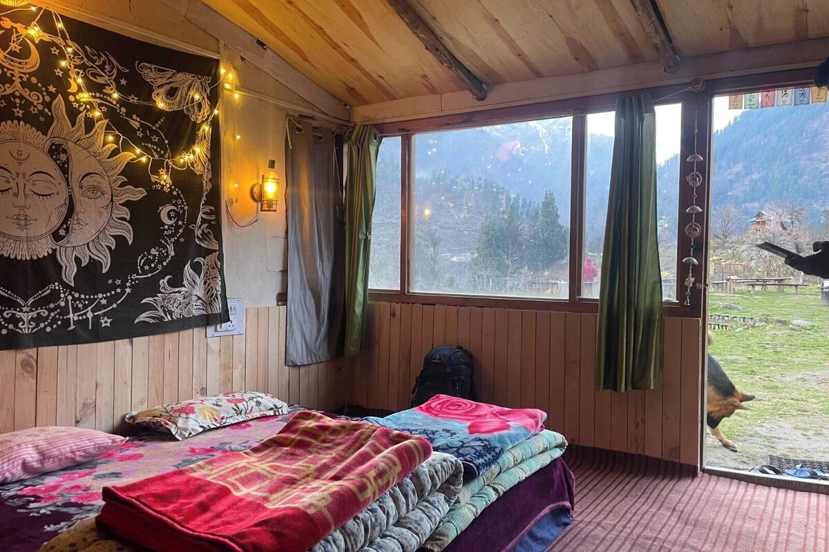 A cozy room with wooden walls, a large window with curtains, a tapestry, and a bed with multiple blankets.
