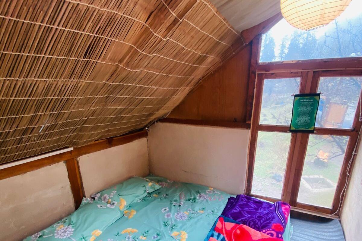 A small, cozy room with a sloped ceiling, a bed with floral and checkered bedding, a hanging light fixture, and a view through a window.