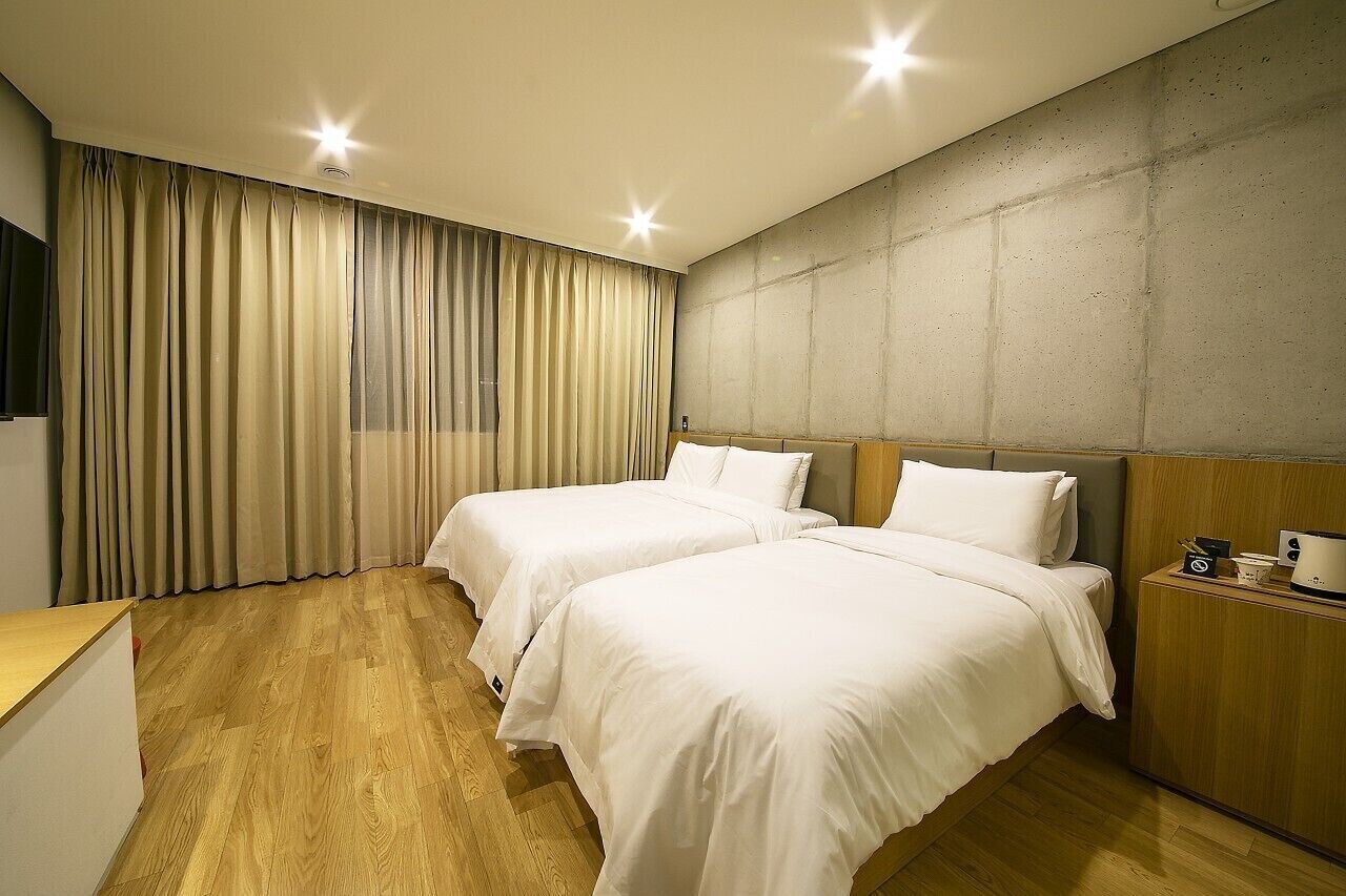 Habitación de hotel con dos camas, suelo de madera, un ventanal con cortinas, pared de acento de concreto y una mesita de noche con lámpara.