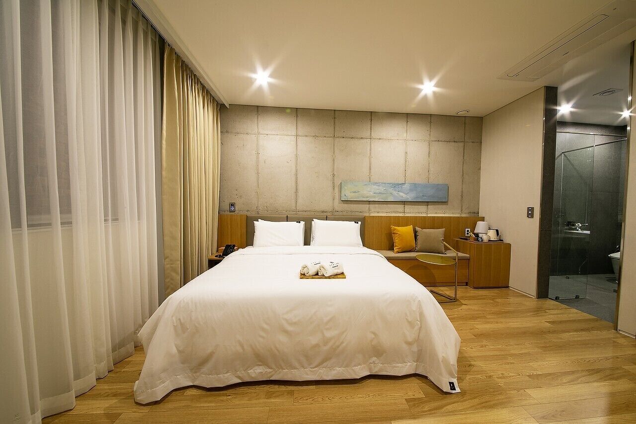 A modern hotel room with a large bed, a bedside table with a lamp, a glass shower enclosure, and a painting on the wall.