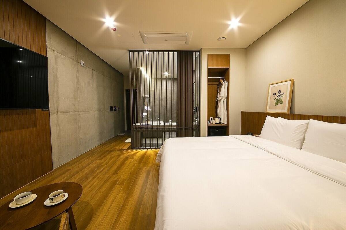A modern hotel room with a large bed, a TV mounted on the wall, a wooden floor, and a closet with a robe and a framed picture.