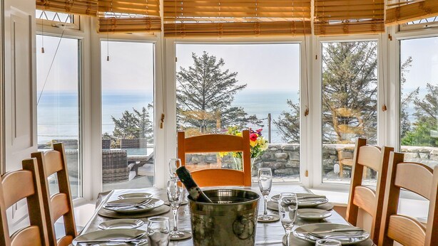 A dining table set for six with a view of the sea through large windows. - Mynydd ar Mor (Barmouth)