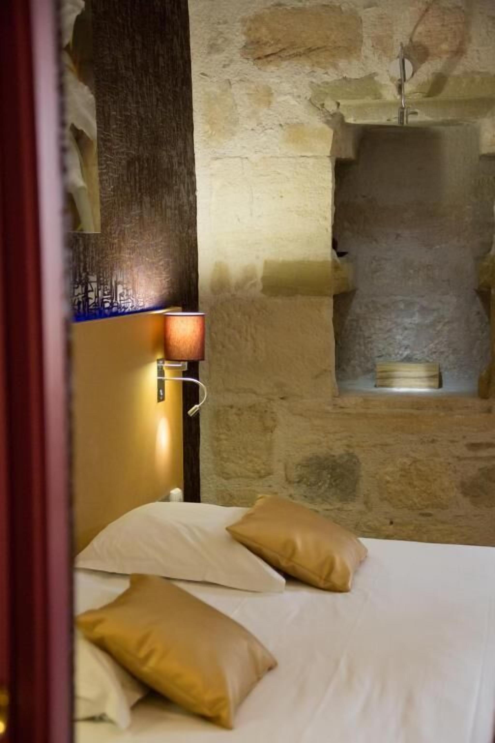 A bedroom with a stone wall, a bed with white and beige pillows, a nightstand with a lamp, and a small shelf with a book.