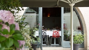 An outdoor dining area with a table set for two, surrounded by potted plants and a glass door leading to an interior space. - Hôtel Le Turenne (Beaulieu-sur-Dordogne)