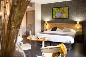 Habitación de hotel con una cama grande, una mesa redonda con una silla, una mesita con una jarra y vasos, y un cuadro de flores colgado en la pared.