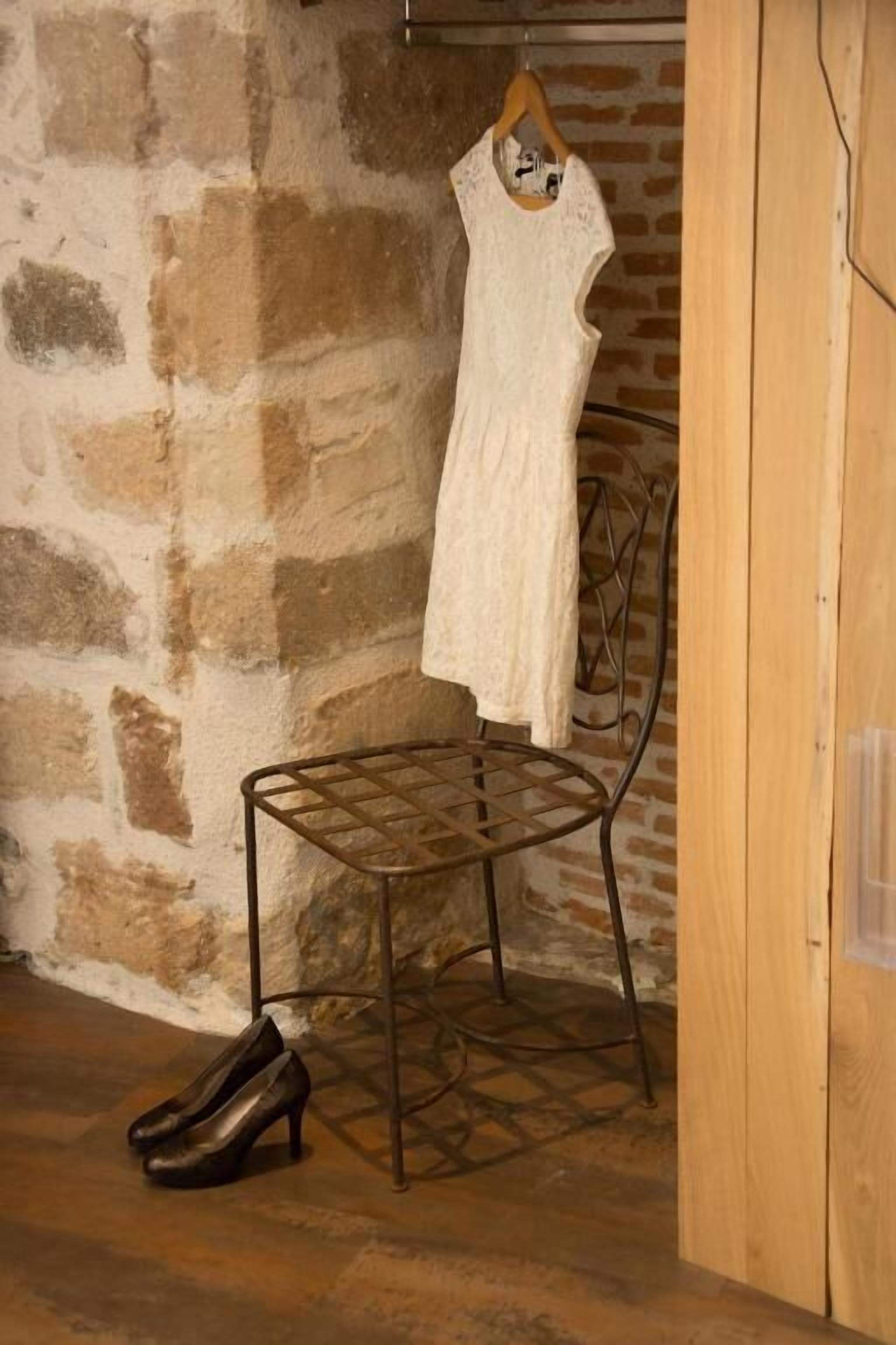 A room with a stone wall, a metal chair, a wooden cabinet, and a white dress hanging on a rack.