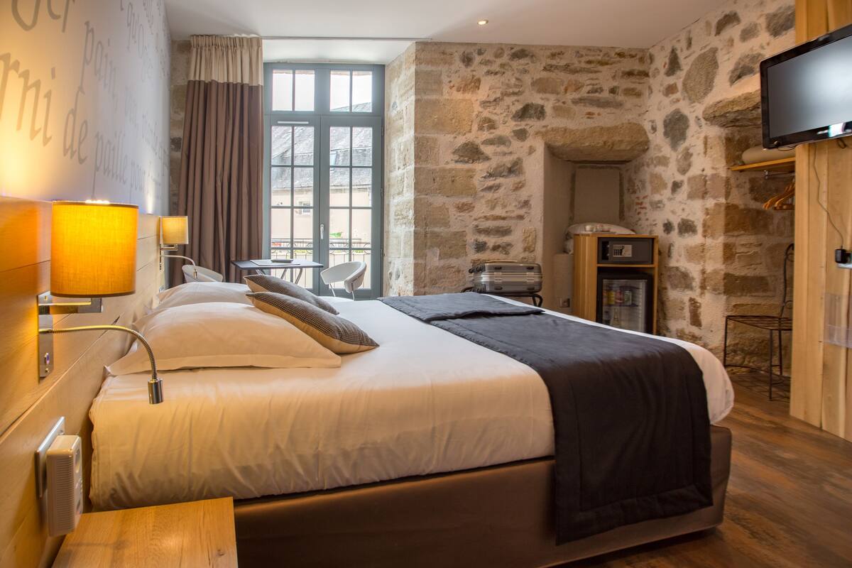 A modern hotel room with a stone wall, a large bed with white and dark bedding, a flat-screen TV, and a wooden nightstand.