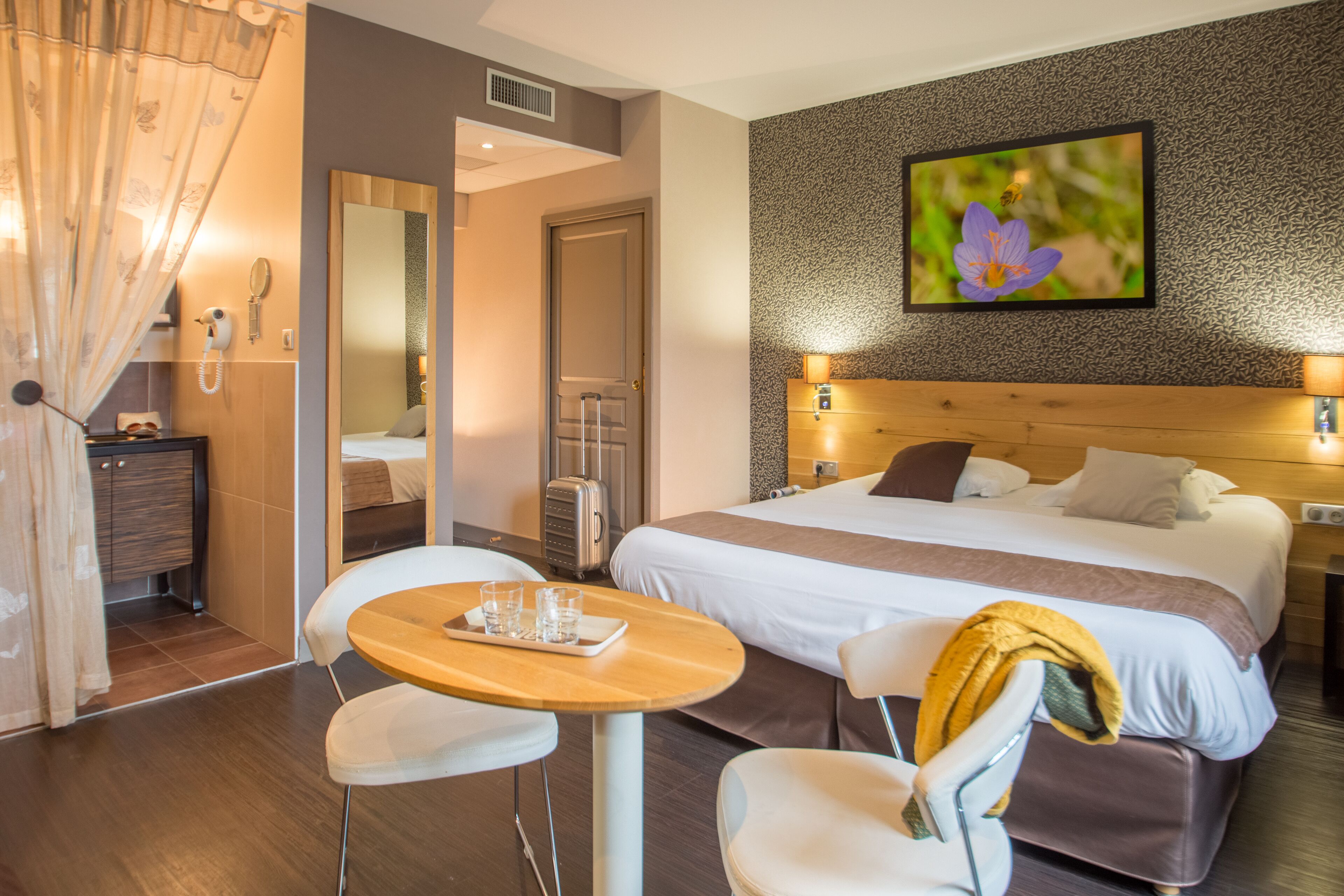 Una habitación de hotel con cama, mesitas de noche, un escritorio con sillas, un espejo y un cuadro en la pared.