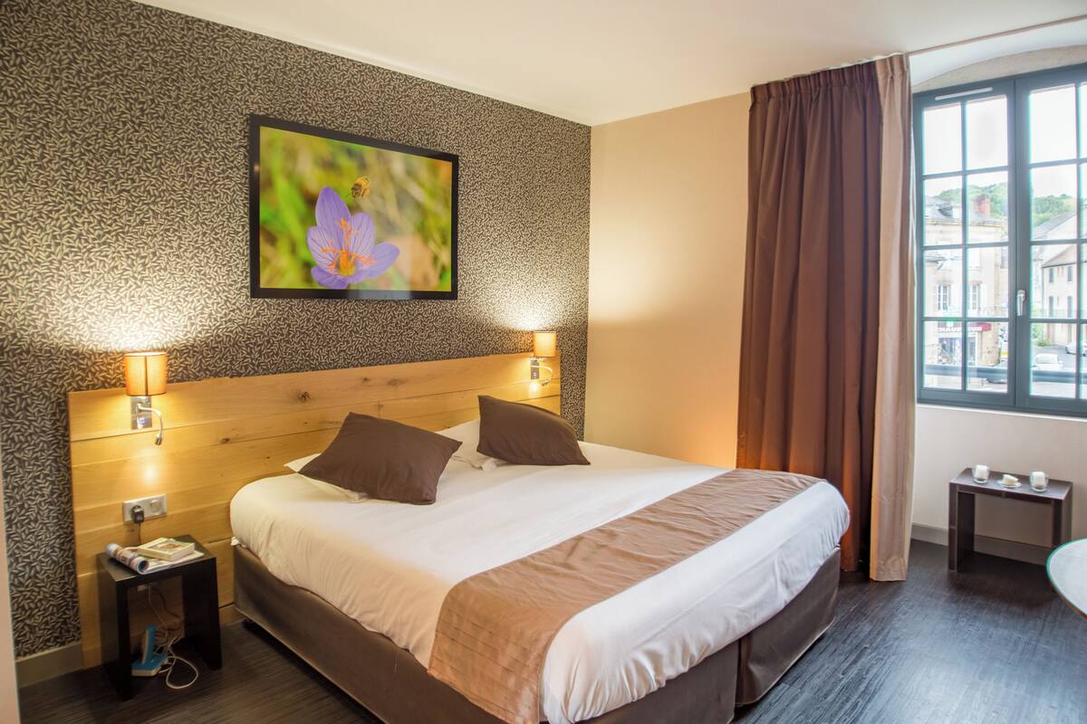 A hotel room with a large bed, two bedside lamps, a nightstand, a small table, a window with curtains, and a framed picture on the wall.