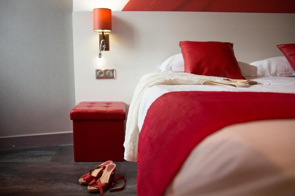 A hotel room with a bed, red pillows, a red ottoman, and a red wall lamp.