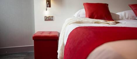 A hotel room with a bed, red pillows, a red ottoman, and a red wall lamp.