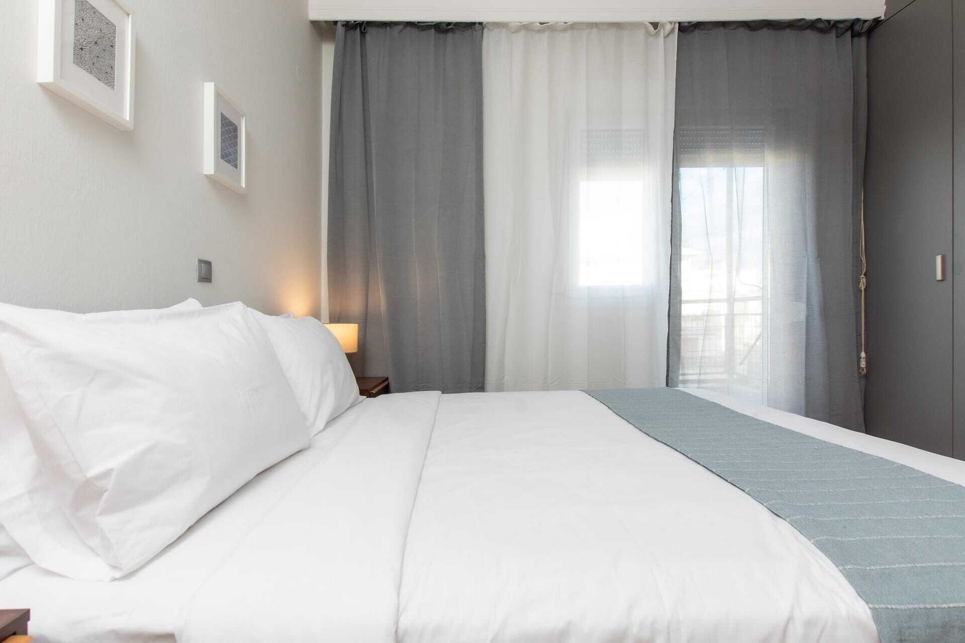Un lit bien fait, avec du linge de lit blanc, une couverture grise et un rideau gris partiellement tiré sur le côté.