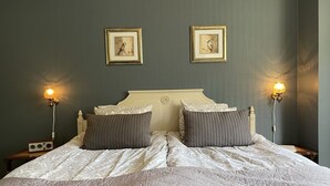 A neatly made bed with a white and grey patterned duvet, two grey pillows, and a headboard with a decorative crest. There are two wall-mounted picture frames with artwork, and two bedside lamps with white shades. - AlingsÃ¥s Vandrarhem - Villa Plantaget - Hostel (Alingsas)