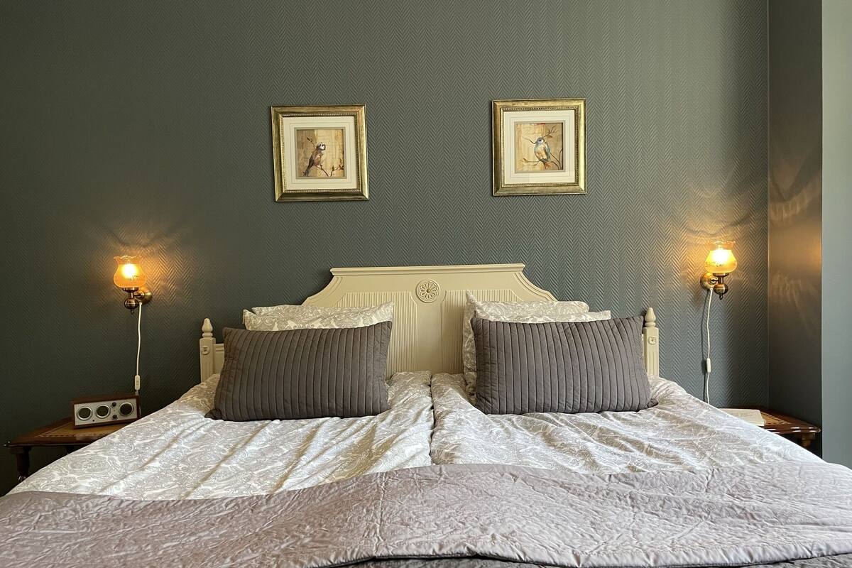 Ein ordentlich bezogenes Bett mit einer weißen und grauen gesteppten Decke, zwei grauen Kissen und einem Kopfteil mit einem dekorativen Wappen. An der Wand hängen zwei Bilderrahmen mit Kunstwerken, und auf den Nachttischen stehen je eine Nachttischlampe mit weißen Schirm