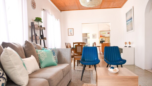 Ein Wohnzimmer mit einer Couch, zwei blauen Sesseln, einem Couchtisch und einem Essbereich mit Stühlen und einem Tisch.