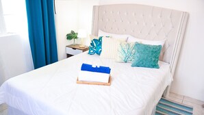 Ein ordentlich gemachtes Bett mit weißer Bettdecke und dekorativen Kissen, ein Nachttisch mit Lampe sowie ein blauer Vorhang.