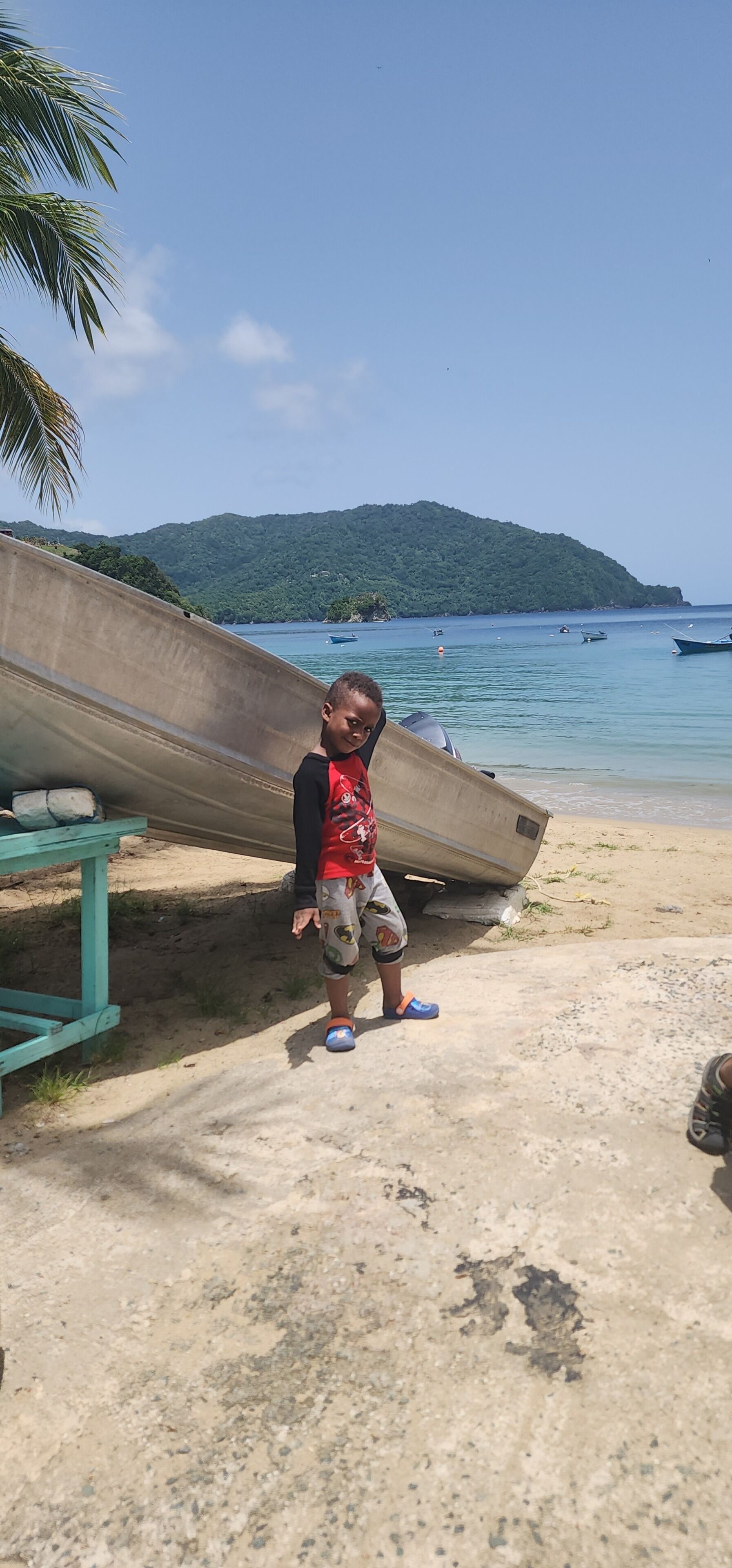 A beach scene with a concrete structure, a child standing next to it, and boats in the water.