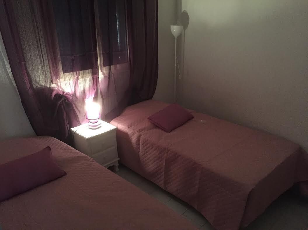 A small, dimly lit room with two single beds, a nightstand with a lamp, and a window with curtains.