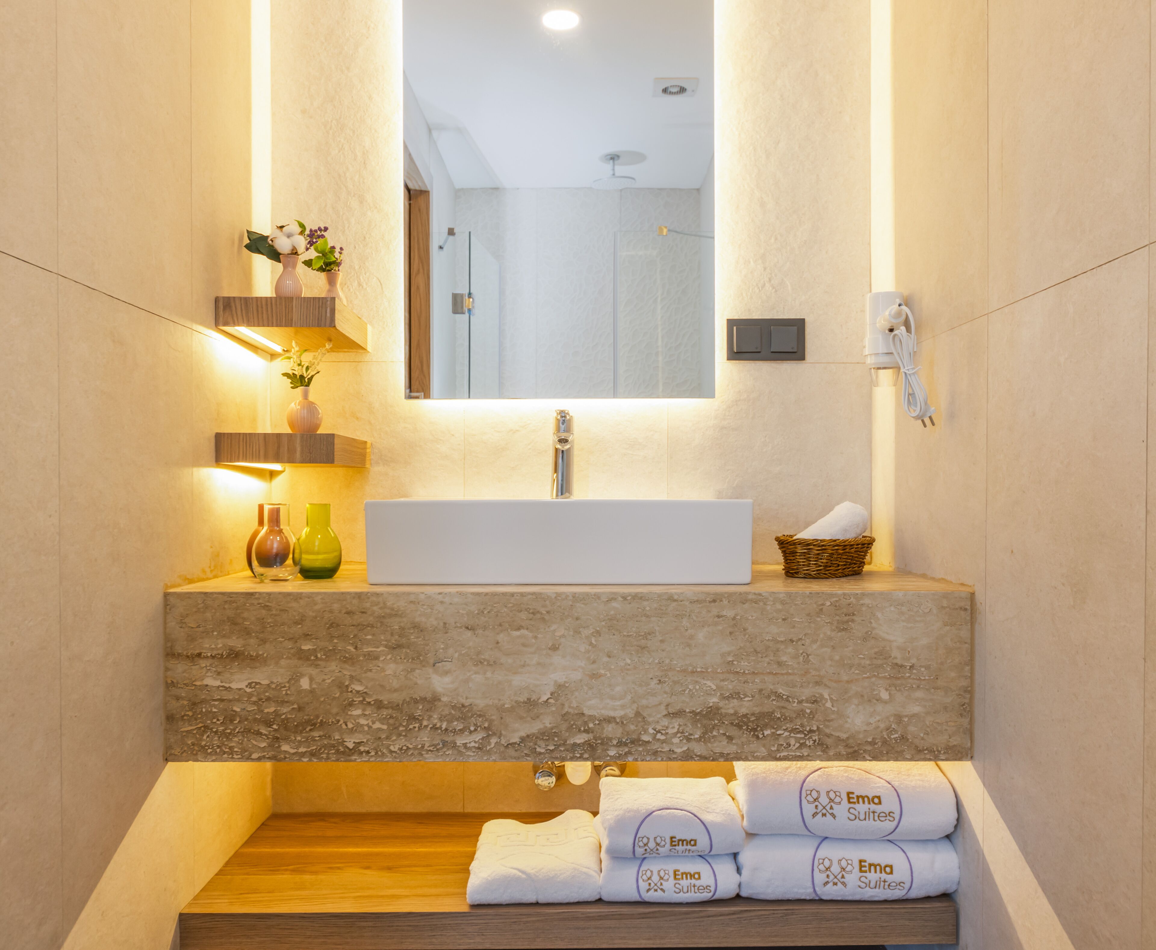 A modern bathroom with a large mirror, a white sink, and a wooden shelf with decorative items. There are towels stacked on the floor and a basket with a towel on the sink.