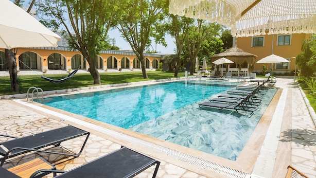 A swimming pool with lounge chairs and umbrellas, surrounded by trees and a building with arches.