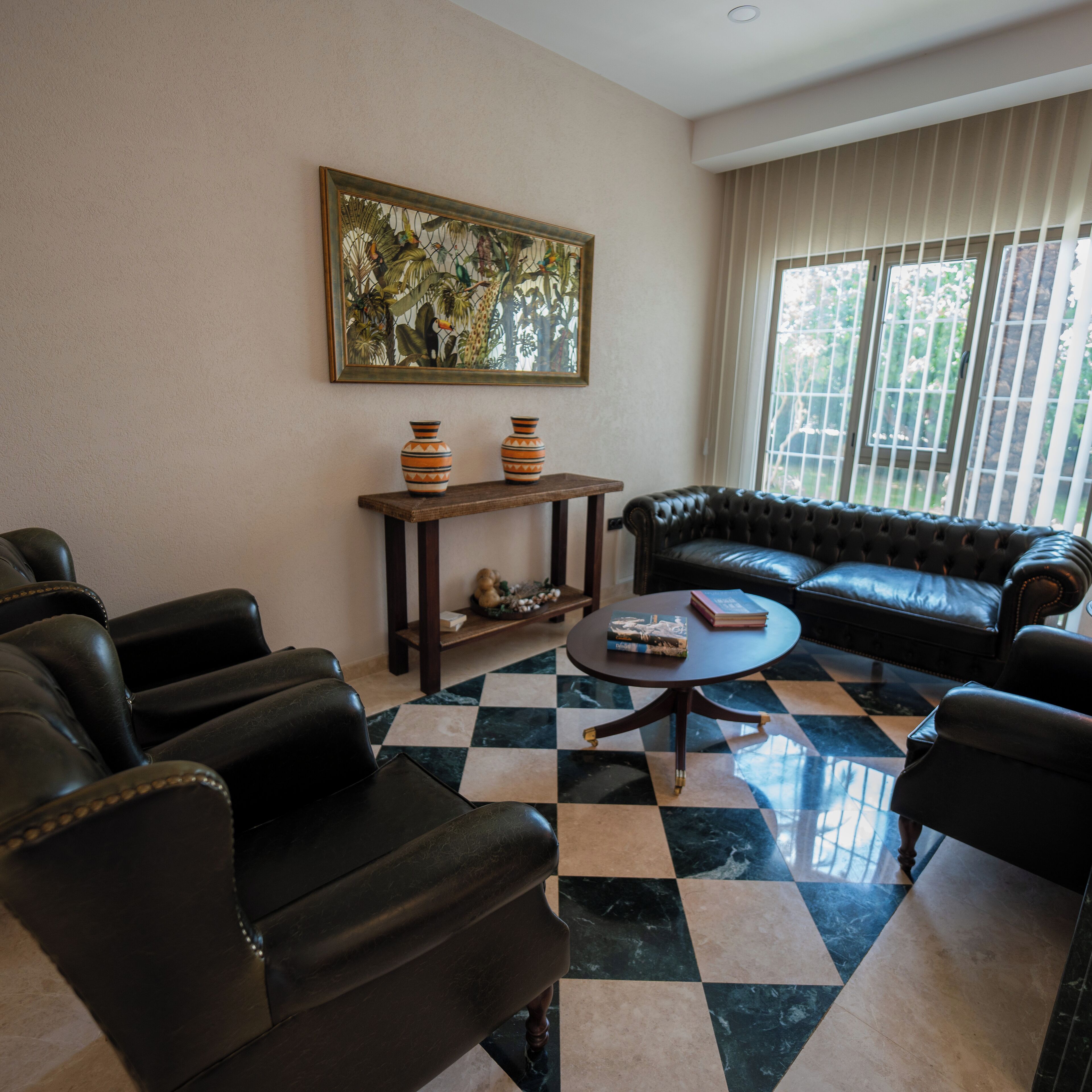 A living room with a checkered floor, a black leather sofa, and two armchairs. There is a small round table with books and decorative items. A painting hangs on the wall above a wooden console table with two vases.