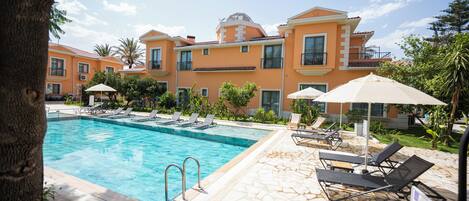 Une piscine entourée de chaises longues et de parasols, elle-même bordée d’un bâtiment doté de balcons et d’un toit en tuiles.