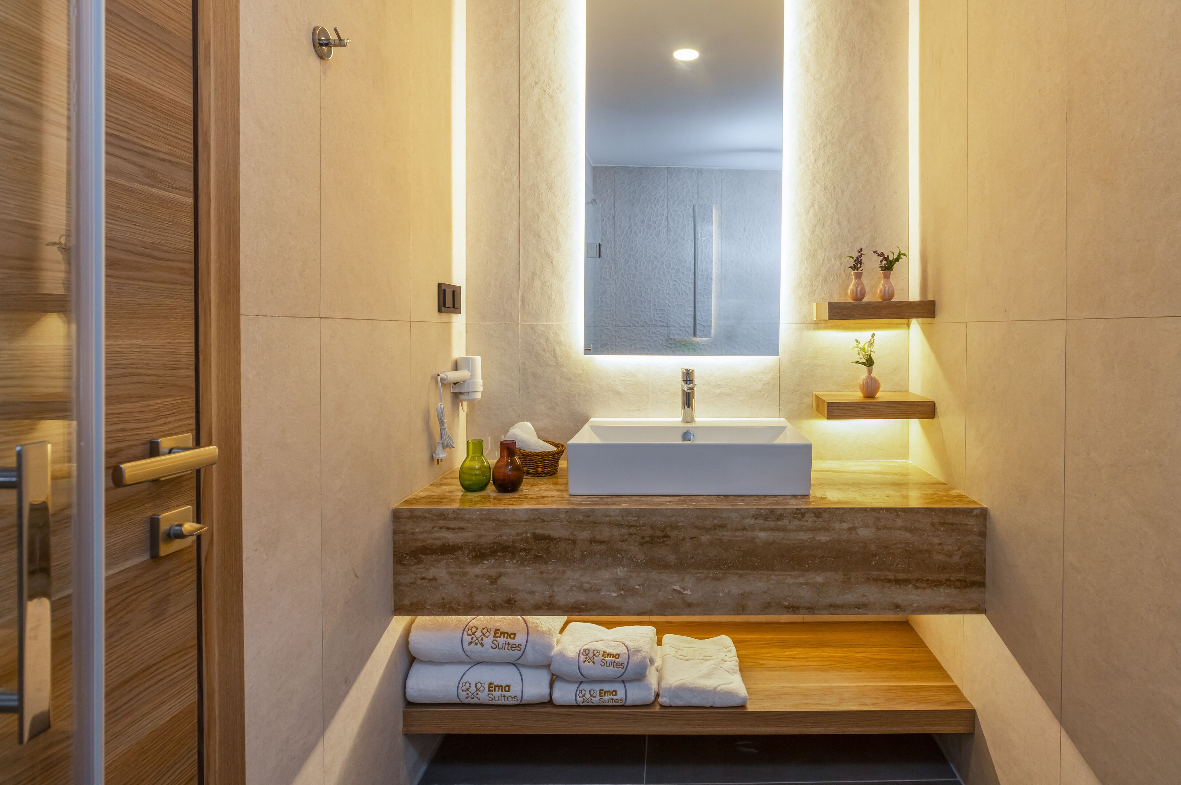 A modern bathroom with a wooden vanity, a large mirror, and a built-in sink. There are towels and toiletries on the vanity, and a shelf with decorative items above the sink.