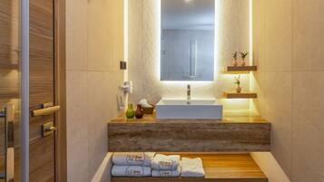 A modern bathroom with a wooden vanity, a large mirror, and a built-in sink. There are towels and toiletries on the vanity, and a shelf with decorative items above the sink.
