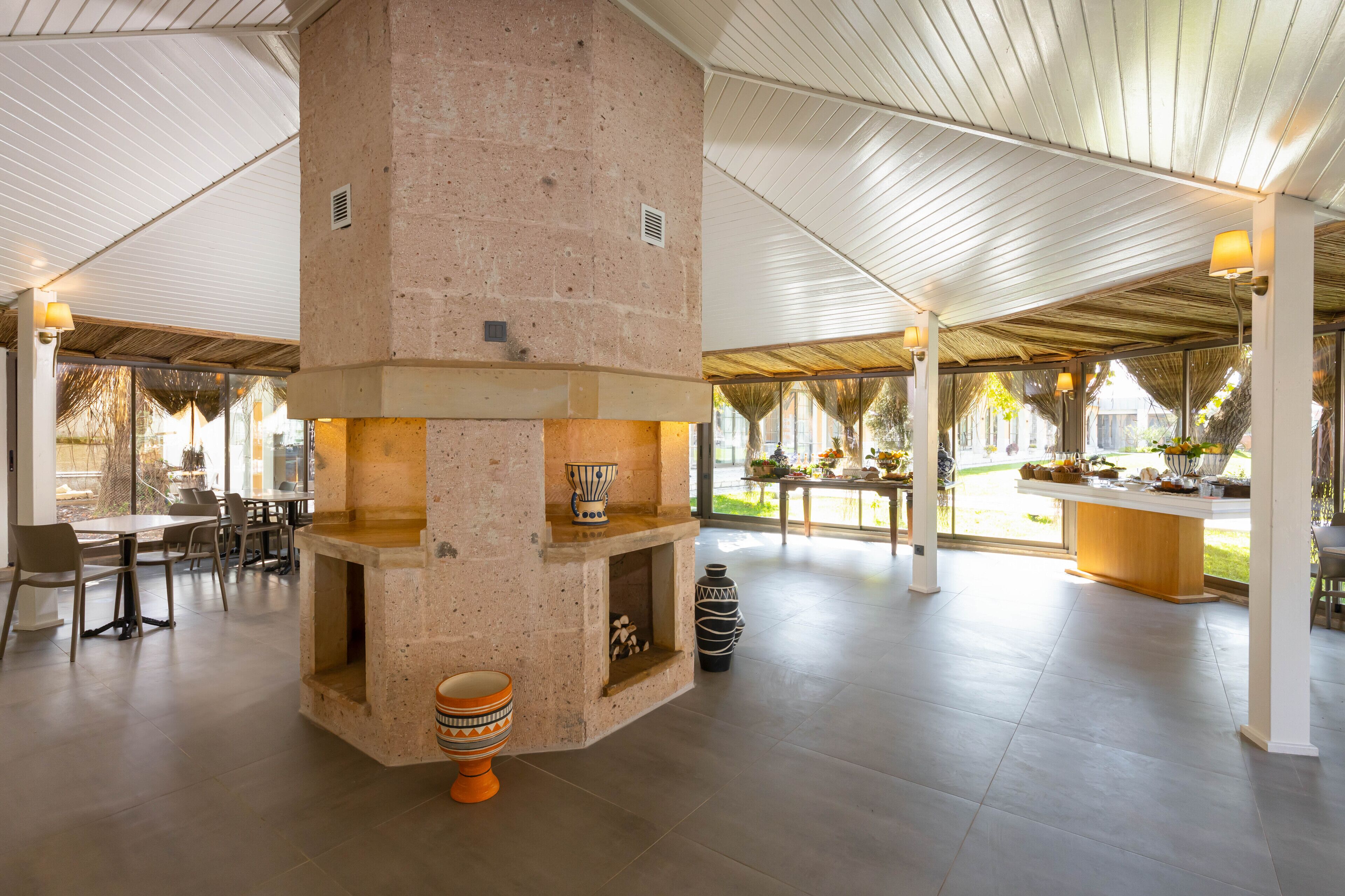 A spacious indoor area with a central stone fireplace, seating arrangements, and a view of the outdoors.
