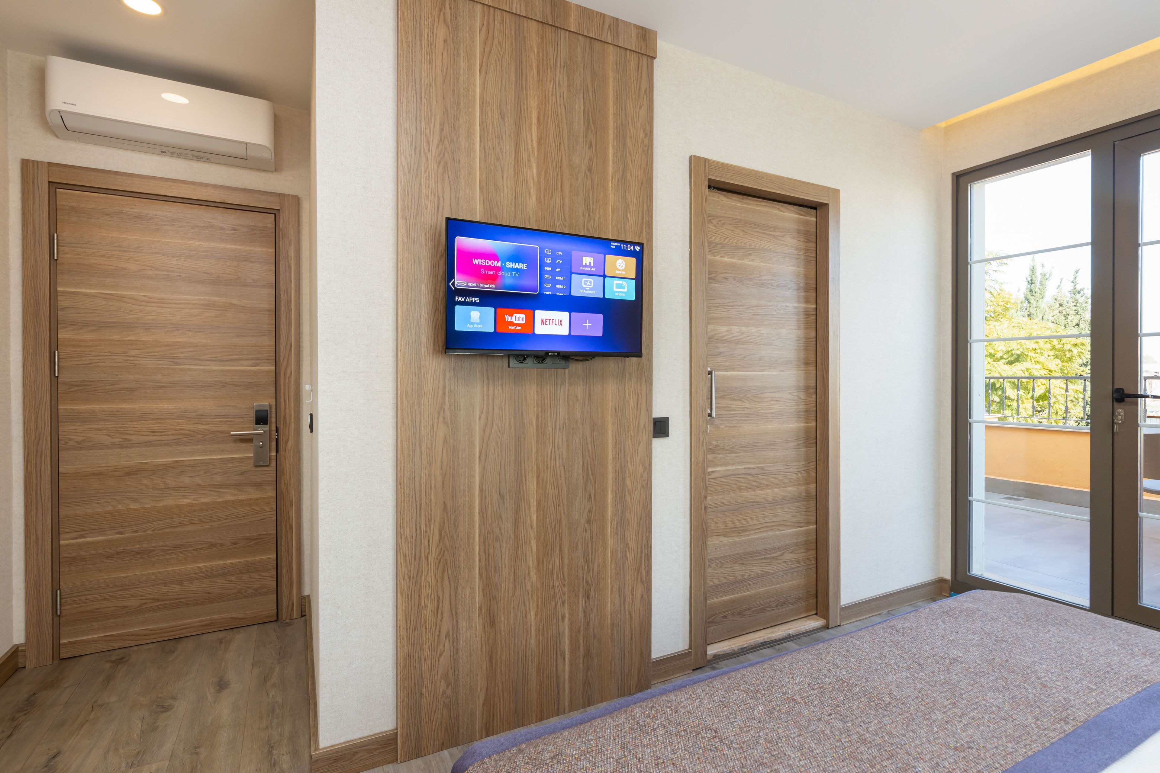 Habitación de hotel con televisor de pantalla plana en la pared, puertas de madera y vistas al exterior a través de un ventanal.
