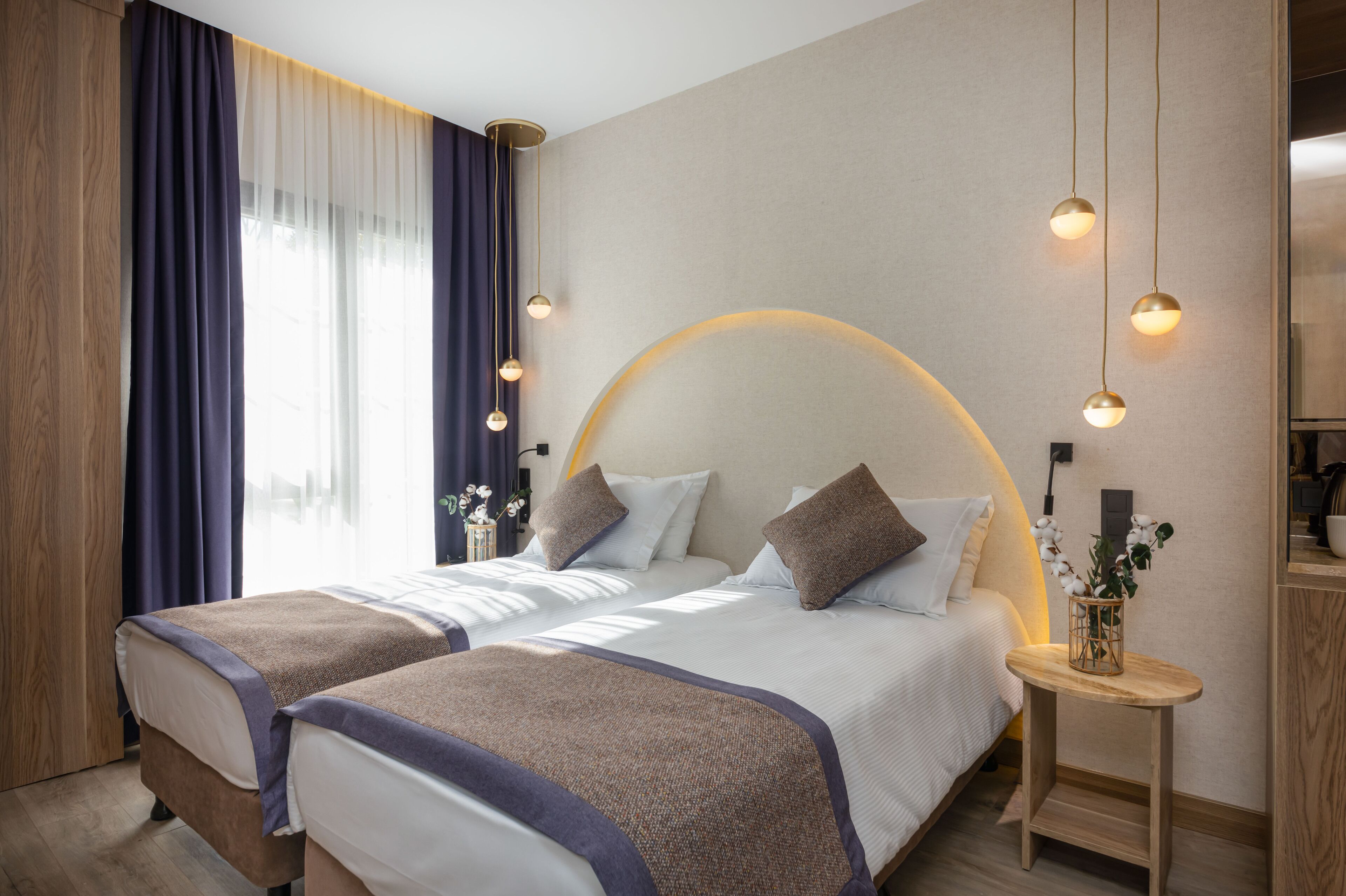 A modern hotel room with two beds, a wooden wardrobe, a round wall light, and a small table with a vase.