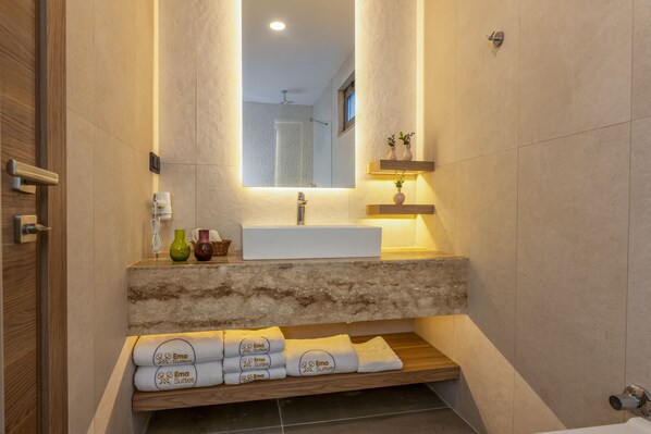 A modern bathroom with a large mirror, a white sink, and a wooden shelf with towels and decorative items.