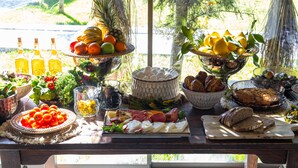 A table set with various food items, including fruits, cheeses, and breads, arranged on a wooden table with a view of greenery outside.
