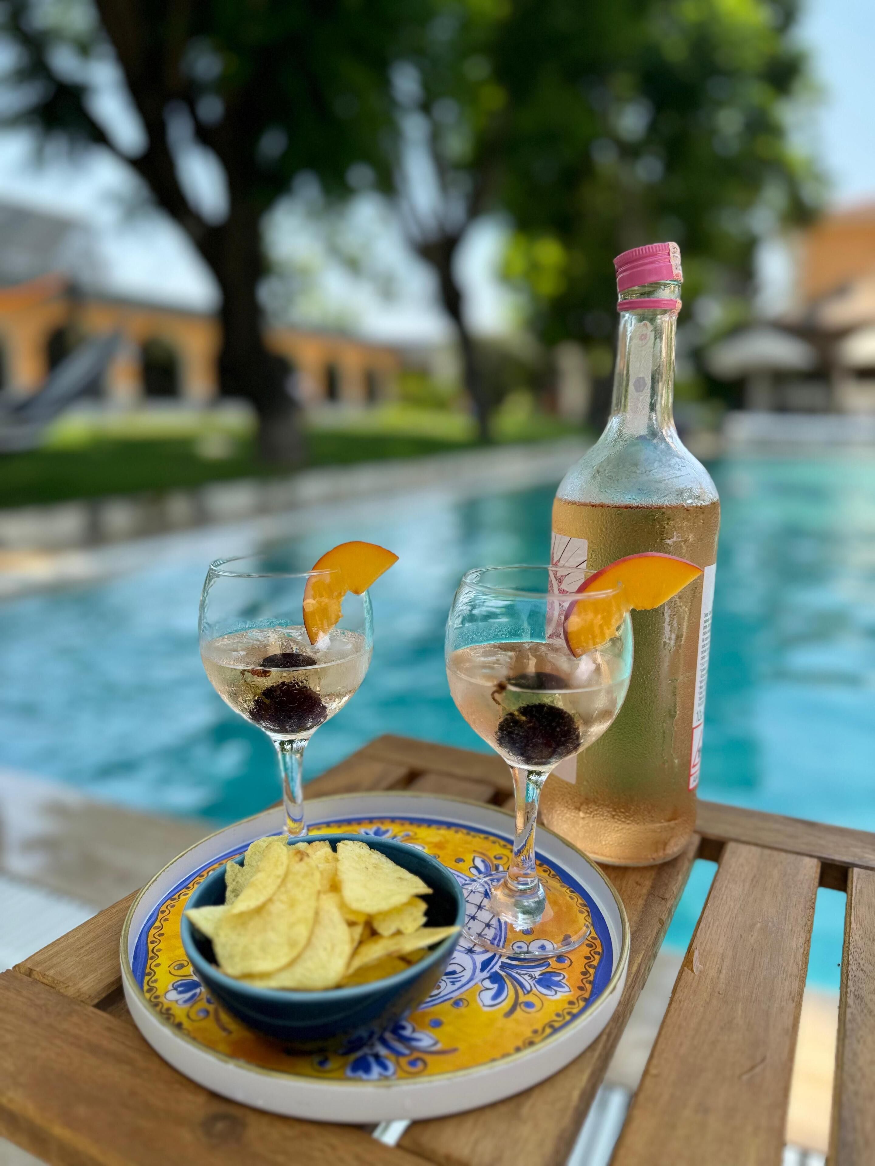 Une table avec deux verres contenant une boisson claire, une bouteille et un bol de chips, près d’une piscine.