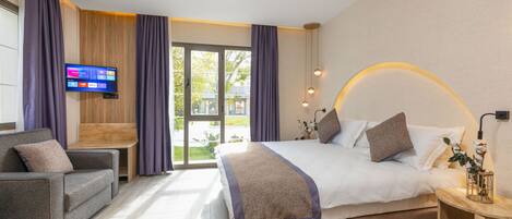 A modern hotel room with a large bed, a sofa, a TV, and a view of the outdoors.