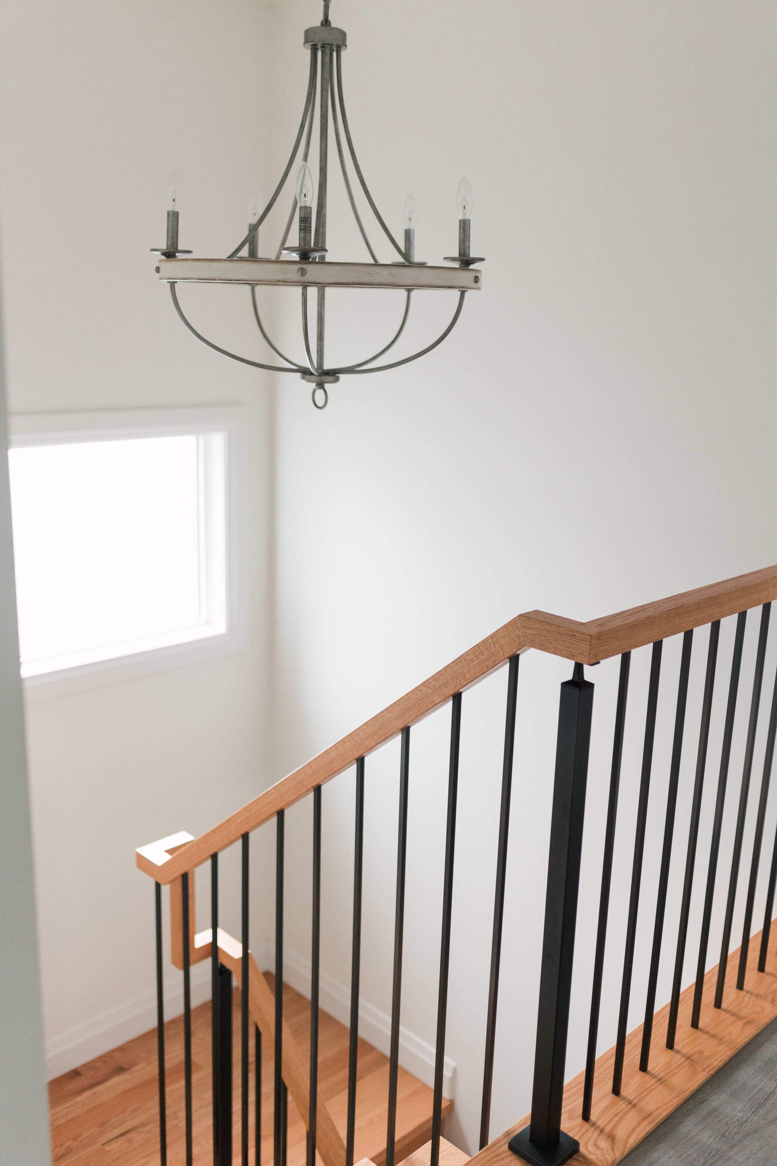 Un escalier doté d’une main courante en bois et de balustrades en métal noir, menant à une pièce avec un mur blanc et une fenêtre.