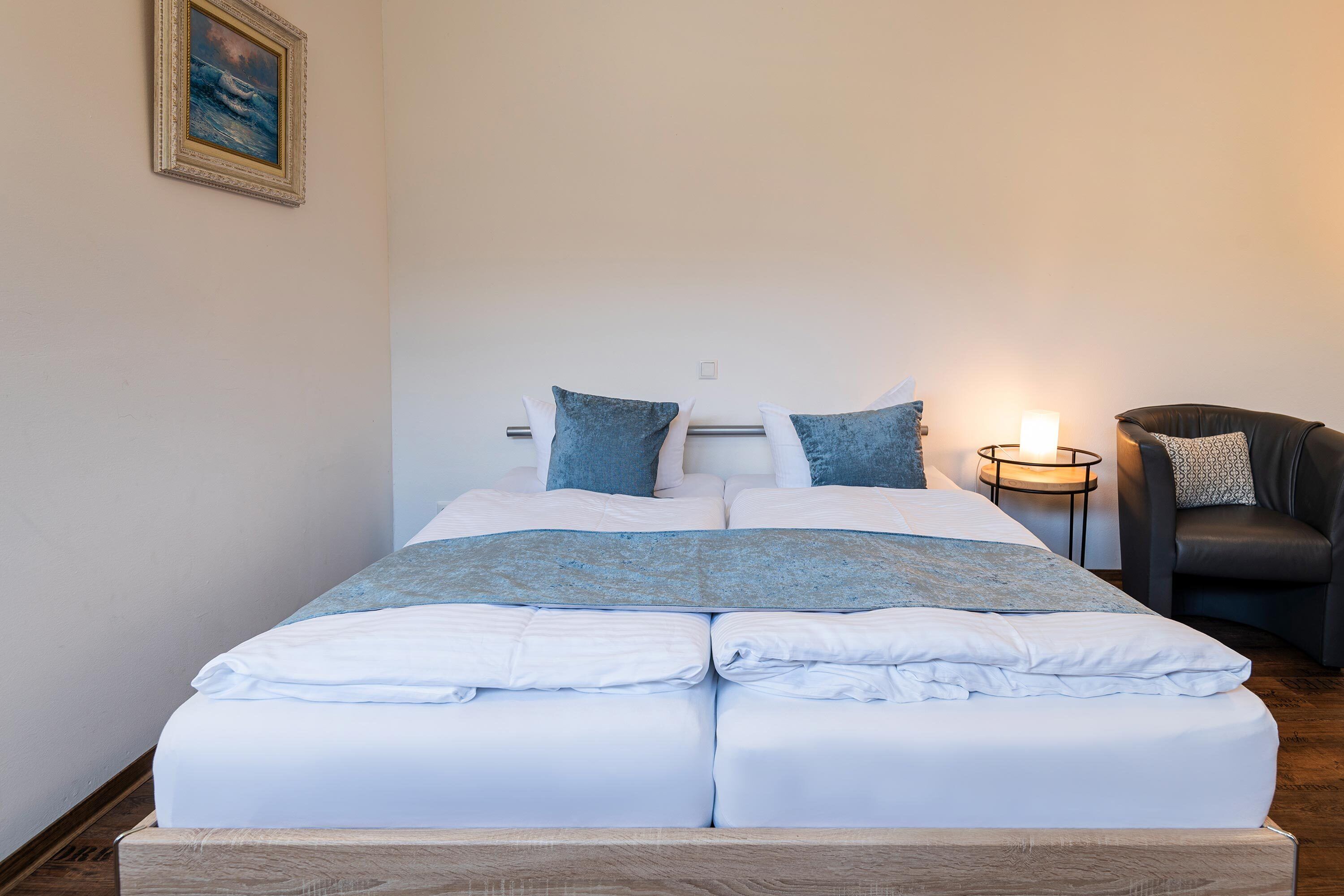 Een net opgemaakt bed met witte lakens en blauwe accenten, een nachtkastje met een kaars, en een stoel met een gestreept kussen.