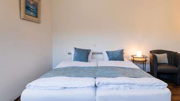Een net opgemaakt bed met witte lakens en blauwe accenten, een nachtkastje met een kaars, en een stoel met een gestreept kussen.