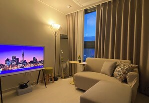 Moderni olohuone, jossa on seinälle asennettu taulutelevisio, joka näyttää kaupunkimaisemaa, sohva, pieni pöytä ja lattiavalaisin.